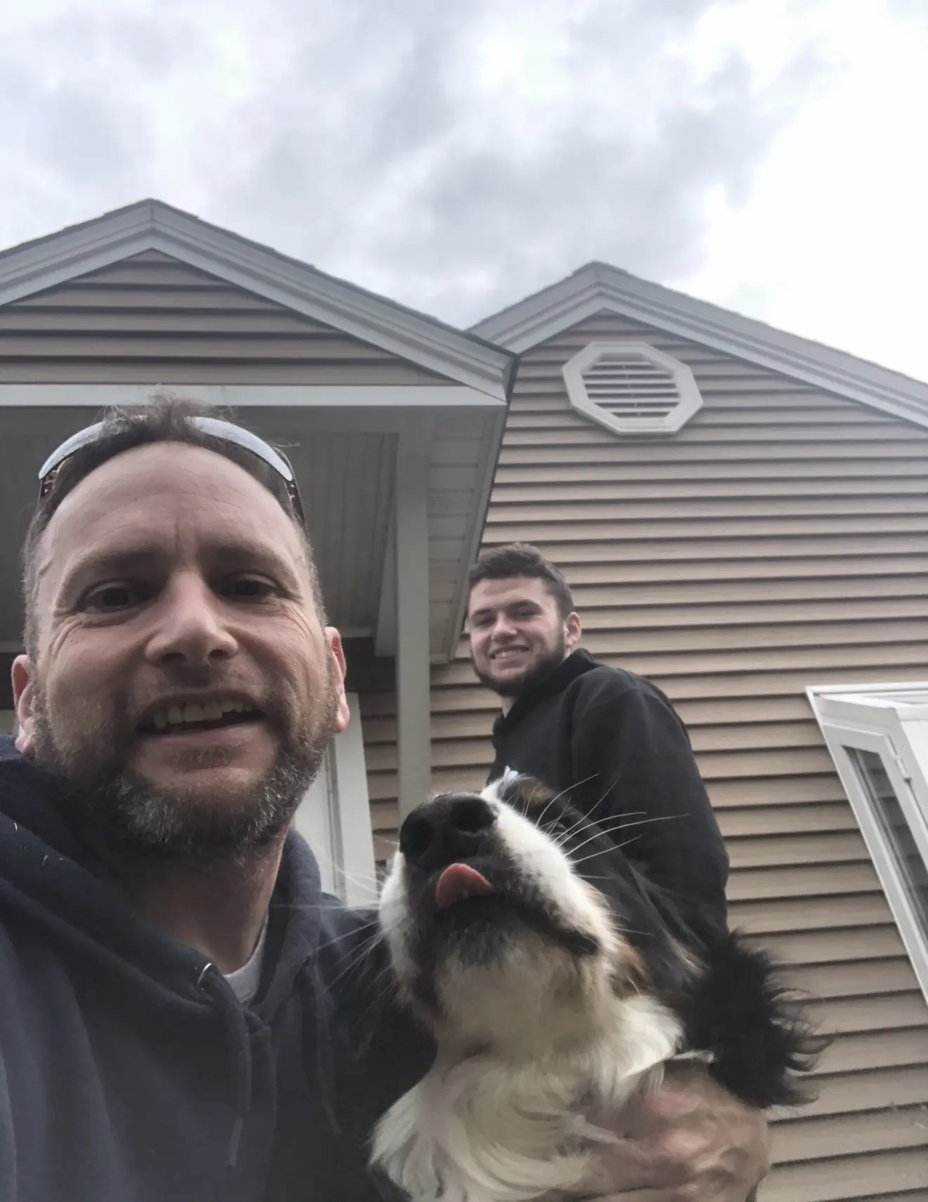 Two men and a dog pose for a selfie in front of a house with cloudy skies above.