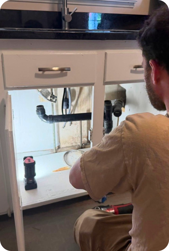 A person inspects plumbing under a kitchen sink, using a flashlight and tools for maintenance.