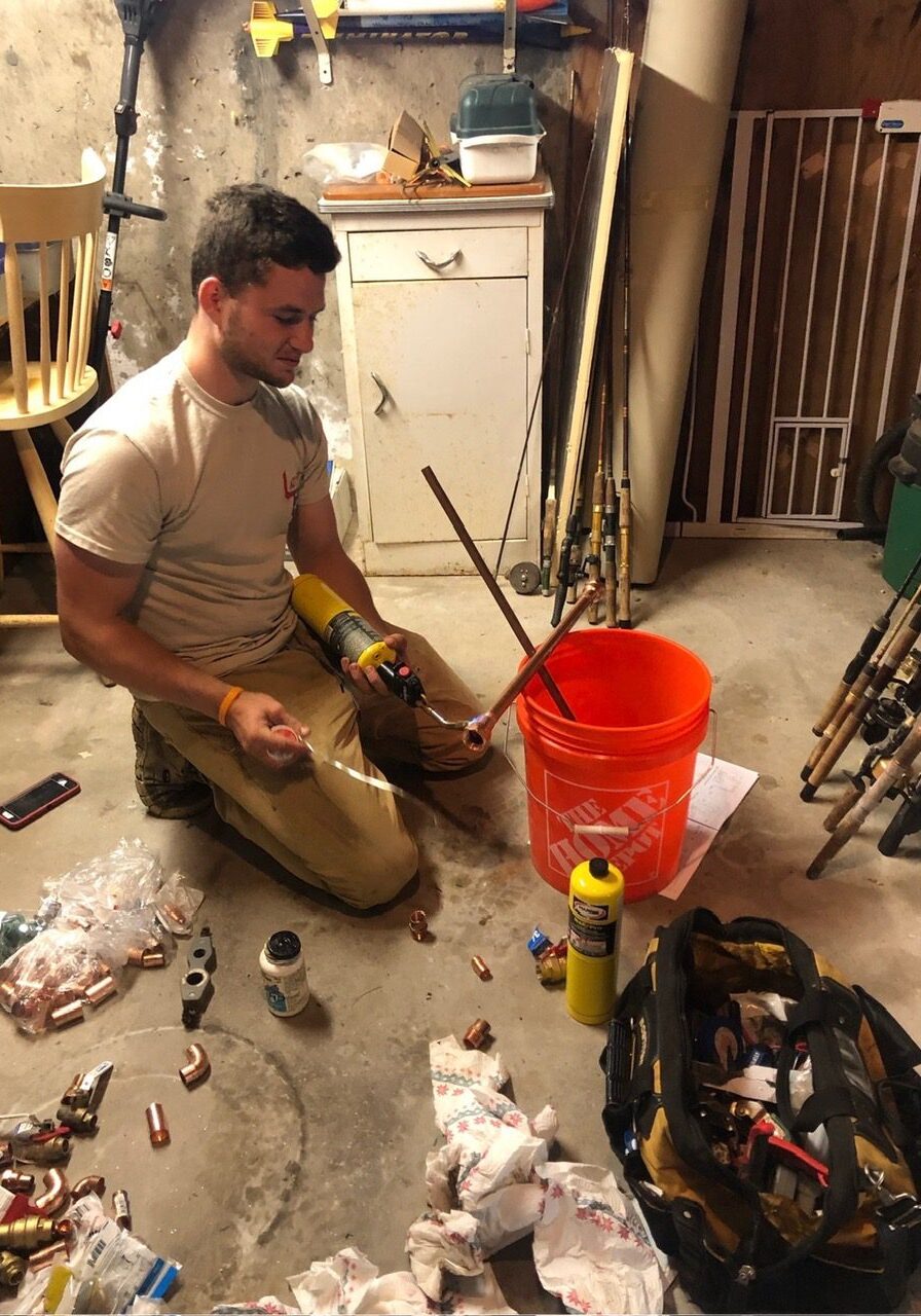 A young man works on plumbing projects in a cluttered basement, surrounded by tools and materials.